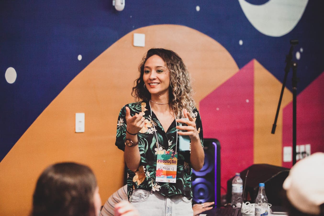 Hellora apresentando em um evento, sorrindo e gesticulando, com um mural colorido ao fundo